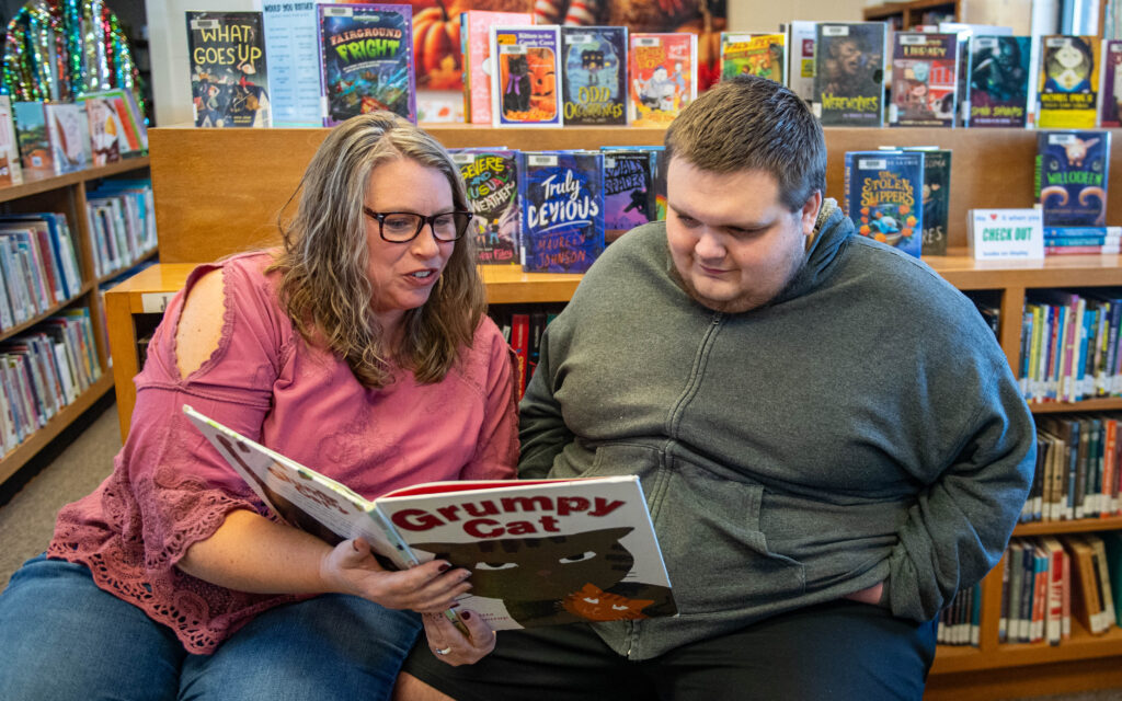 A woman is reading to a young man in a library.